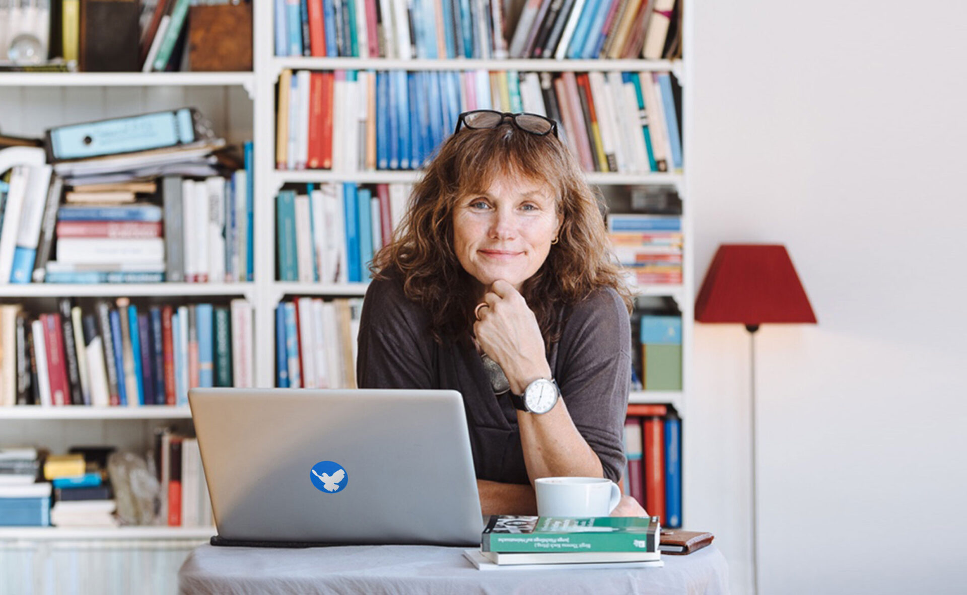 Portrait von Birgit Theresa Koch, sie sitzt am Schreibtisch vor ihrem aufgeklappten Laptop. Den Kopf auf die Hand abgelegt, lächelt sie frontal in die Kamera.